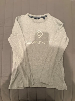 Grå långärmad topp från Gant - Snygg grå långärmad topp från Gant med rund halsringning och vit logga framtill. Tillverkad i mjuk bomull som känns skön mot huden. Perfekt basplagg med enkel och stilren design.