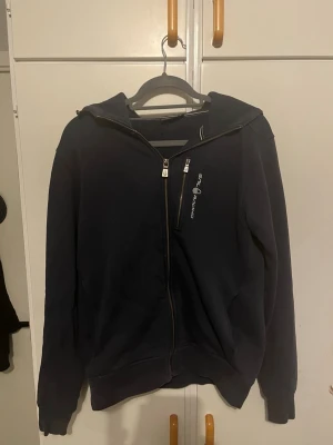 Svart zip hoodie från Sail Racing - Snygg svart hoodie från Sail Racing med dragkedja och huva. Har en diagonal dragkedja upptill och logga tryckt på bröstet. Tillverkad i mjukt bomullsmaterial och har ribbade muddar vid ärmar och nederkant. Perfekt för en avslappnad streetwear-look.