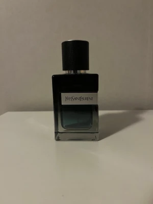 Yves Saint Laurent parfymflaska - Snygg parfym från Yves Saint Laurent i en stilren, fyrkantig glasflaska med svart lock. Flaskan har en mörk toning från svart till transparent och en metallisk etikett med loggan på framsidan. Perfekt för dig som gillar exklusiva dofter.