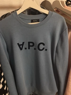 Blå sweatshirt från A.P.C. - Snygg blå sweatshirt från A.P.C. med svart logotyp tryckt framtill. Tröjan har rund halsringning och långa ärmar. Tillverkad i mjukt material som känns skönt mot huden. Perfekt för en avslappnad och stilren look.