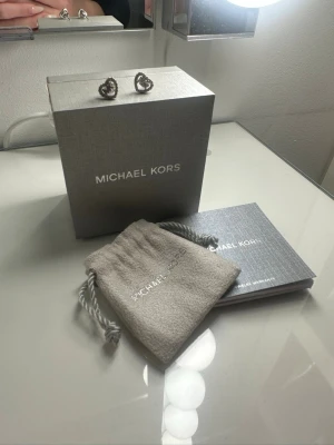 Silverfärgade hjärtörhängen Michael Kors - Snygga örhängen från Michael Kors i form av hjärtan. De är silverfärgade och har glittrande stenar runt hela hjärtat för extra bling. Perfekta för dig som vill ha en elegant och trendig accessoar. Kommer med originalask och tygpåse.
