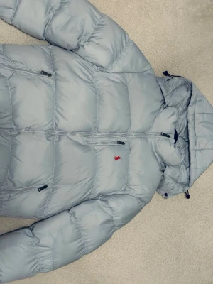 Ljusgrå dunjacka från Polo Ralph Lauren - Snygg ljusgrå dunjacka från Polo Ralph Lauren med röd broderad logga på bröstet. Jackan har huva, dragkedja framtill och två sidofickor med dragkedja. Perfekt för kalla vinterdagar och riktigt skön att bära.