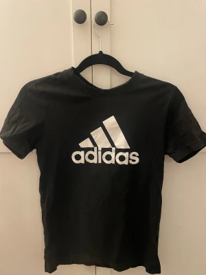 Svart Adidas t-shirt med logga - Snygg svart t-shirt från Adidas med den klassiska vita loggan tryckt framtill. T-shirten har rund halsringning och korta ärmar. Tillverkad i mjuk bomull som känns skön mot huden. Perfekt för en sportig och avslappnad stil.🥰💕