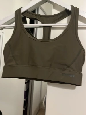 Olivgrön sport-bh från Stronger - Snygg olivgrön sport-bh från Stronger med brottarrygg. Tillverkad i ett stretchigt material av 75% återvunnen polyester och 25% elastan. Perfekt för träning eller som ett bekvämt basplagg. Diskret logga framtill.