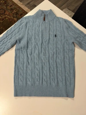 Ljusblå kabelstickad tröja Polo Ralph Lauren - Ljusblå kabelstickad tröja från Polo Ralph Lauren med halv dragkedja och hög krage. Tröjan har ribbade muddar och nederkant samt den klassiska broderade loggan på bröstet. Perfekt för lager på lager och riktigt snygg till jeans.