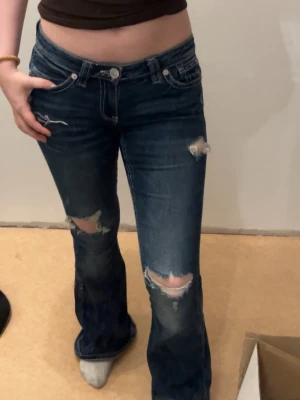 Mörkblå bootcut jeans med slitningar - Snygga mörkblå bootcut jeans med slitna detaljer och hål på knäna. Jeansen har klassisk femficksdesign, låg midja och dragkedja med knapp. Perfekta för en avslappnad och trendig look.