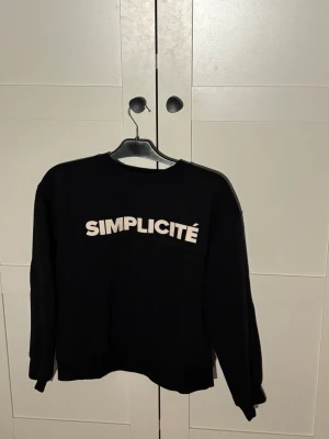 Svart sweatshirt med SIMPLICITÉ-tryck - Svart sweatshirt med vitt SIMPLICITÉ-tryck över bröstet. Tröjan har rund halsringning och långa ärmar. Enkel och stilren design som passar perfekt till jeans eller joggers. Mjuk och bekväm bomullskänsla.