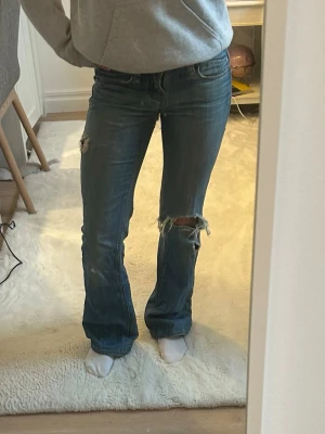 Blå bootcut jeans med slitningar - Snygga blå bootcut jeans med slitningar på knäna och låret. Jeansen har klassisk femficksdesign och detaljerade bakfickor med mönster. Hål i skrevet som lätt går att sy ihop! Är osäker på storleken men uppskattar den till XS