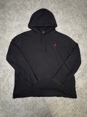 Svart hoodie från Polo Ralph Lauren - Svart hoodie från Polo Ralph Lauren med klassisk röd broderad logga på bröstet. Hoodien har huva med dragsko och långa ärmar. Tillverkad i mjukt bomullsmaterial som känns skönt mot huden. Perfekt för en avslappnad och stilren look. Hoodien har dock en lite skroma så är ordentligt lagat😊