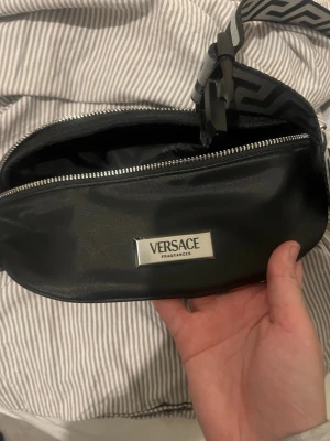 Svart magväska från Versace - Snygg svart magväska från Versace med silverfärgad logga framtill. Väskan har ett stort fack med dragkedja och ett justerbart band med mönster i svart och grått. Perfekt för att bära det viktigaste och ger en trendig touch till din outfit.