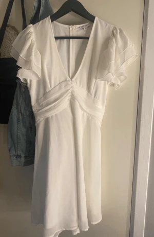 Vit kortklänning med volangärm  - Vit klänning med v-ringning och volangärmar. Klänningen har omlott detaljer över bysten och dragkedja i ryggen. Använd endast en gång. Tveka inte på att höra av dig om du har funderingar kring något!🫶🏻