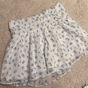 Vit kjol med blå blommor - Söt kort kjol i vitt med blått blommönster. Kjolen har en lätt utsvängd form och är gjord i ett tunt, luftigt material med resår i midjan. Aldrig använd, från Gina tricot Young stl 146/152