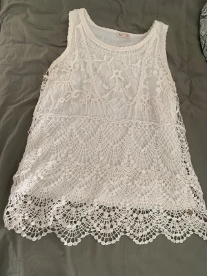 Vit spetslinne från Ü - Säljer ett superfint vitt spetslinne från Ü i storlek 158/164. Linnet har ett detaljerat spetsmönster med blommiga inslag och vågig kant nertill. Perfekt att styla över ett linne eller bikini för en chill look.