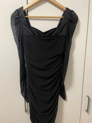 Svart långklänning med meshärmar - Snygg svart långklänning med transparenta meshärmar och rynkade detaljer längs hela klänningen. Klänningen har en rak, figurnära passform och v-ringad urringning. Perfekt för dig som vill sticka ut med en elegant och modern look.