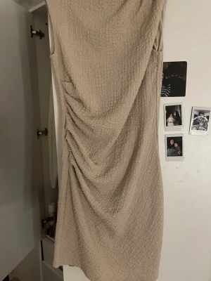 Beige omlott kortklänning - Trendig beige kortklänning med omlottdetalj och snygg rynkning på sidan. Klänningen har en strukturerad yta och är ärmlös, vilket ger en modern och stilren look. Perfekt för dig som gillar minimalistisk stil.