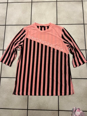 Rosa och svart randig topp från Top-In storlek L - Unik topp i rosa och svart med vertikala ränder och dekorativ snedsydd spetsdetalj framtill. Trekvartsärm och rund halsringning. Perfekt för dig som vill sticka ut med en cool och färgstark stil.