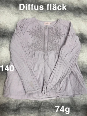 Ljuslila blus med broderi från noanoa  - Ljuslila blus från noanoa med fina broderade detaljer framtill. Blusen har långärmad design, rund halsringning och knäppning framtill. Tillverkad i ett tunt tyg som ger en avslappnad look.🌸Diffus fläck. Storlek 140 