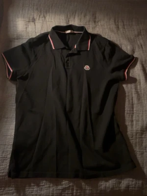 Svart pikétröja från Moncler - Klassisk svart pikétröja från Moncler med korta ärmar och krage. Tröjan har knappar framtill och Moncler-logga på bröstet. Kragen och ärmsluten har tunna ränder i vitt och rött för en sportig look. Perfekt till sommaren.