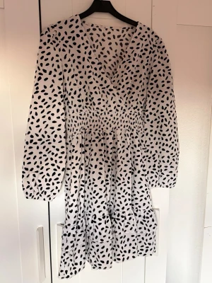 Midiklänning sommar - Snygg vit kortklänning med svarta prickar och v-ringad omlottfront. Klänningen har långa ärmar med resår vid handlederna och smock i midjan för en bekväm passform. Perfekt för dig som gillar trendiga mönster och en luftig känsla.