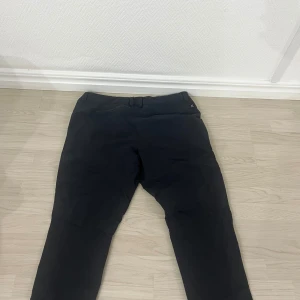 Svarta byxor från Haglöfs - Säljer ett par svarta byxor från Haglöfs med dragkedjor på fickorna och en stilren design. Perfekta för dig som gillar funktionella och snygga byxor med en bekväm passform. Passar till många olika tillfällen och har diskret logga på låret.