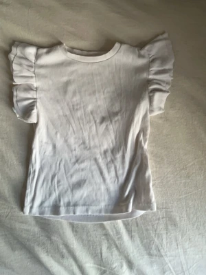 Vit ribbad topp med volangärmar - Supersöt vit ribbad topp med volangdetaljer på ärmarna. Perfekt till jeans eller kjol till våren/sommaren. Tröjan är i st 164 och använd några gånger.