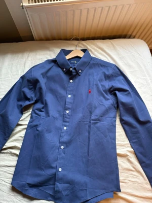 Blå skjorta från Ralph Lauren - Klassisk blå skjorta från Ralph Lauren med button-down krage och röd broderad logga på bröstet. Skjortan har lång ärm, vita knappar och är tillverkad i bomull. Perfekt för en stilren och tidlös look. Använd endast en gång riktigt fin skick 