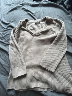 Ljusbrun ribbstickad tröja med v-ringning - Ljusbrun ribbstickad tröja med v-ringad hals och långa ärmar. Tröjan har en avslappnad passform och är tillverkad i ett mjukt stickat material. Perfekt för lager på lager och enkel att matcha med olika stilar. Den har ett hål men det är lätt att fixa om man kan det.