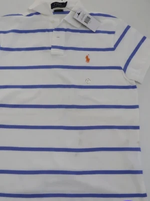 Vit och blå randig piké från Polo Ralph Lauren - Vit pikétröja med blå horisontella ränder från Polo Ralph Lauren. Klassisk krage och korta ärmar, med den ikoniska broderade loggan i orange på bröstet. Perfekt för en avslappnad men stilren look.