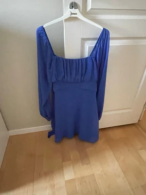 Blå kort klänning med knytband - Supersöt blå klänning med långa ärmar. Klänningen har ett knytband i ryggen och en lätt rynkad överdel som ger en romantisk vibe. Perfekt för sommarens alla tillfällen.