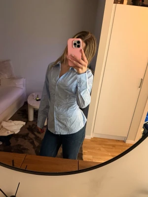 Ljusblå skjorta med snörning bak -  ljusblå skjorta med  långa ärmar. Skjortan har en unik detalj med snörning i ryggen. 