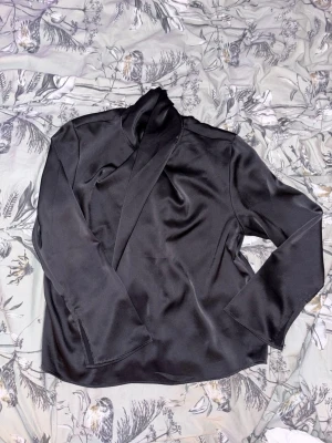 Svart satinskjorta med scarfkrage - Stilren svart blus i glansigt satinmaterial med lång ärm och scarfkrage som har knäppen i nacken. Perfekt för dig som gillar en elegant och trendig look. Passar till både jeans och kjol för en snygg outfit.