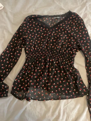 Svart blommig blus från SHEIN - Svart blus från SHEIN med små röda och vita blommor, v-ringning med spetskant och smockad midja. Långa ärmar och lätt, luftig kvalitet som ger en snygg siluett. Perfekt för att lyfta din outfit med en söt touch.