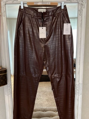 Bruna skinnbyxor från By Malina - Snygga bruna skinnbyxor från By Malina med croco-mönstrad yta. Byxorna har hög midja, bälteshällor och raka ben. Perfekta för dig som vill ha en trendig och edgy look. Stängs med dragkedja och knapp framtill.  Passar M L