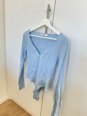 Ljusblå ribbad v-ringad body från NAKD - Säljer en ljusblå ribbad body från NAKD med v-ringning och långa ärmar. Bodyn är tillverkad i mjuk, stretchig bomullsmix. Perfekt för dig som gillar stilrena och bekväma plagg.