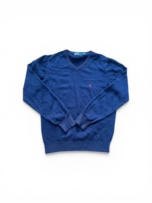 Marinblå v-ringad tröja Polo Ralph Lauren - Klassisk marinblå v-ringad tröja från Polo Ralph Lauren med den ikoniska röda logobrodyr på bröstet. Tröjan har långa ärmar, ribbade muddar och är tillverkad i mjukt material som passar perfekt till lager-på-lager-stil.