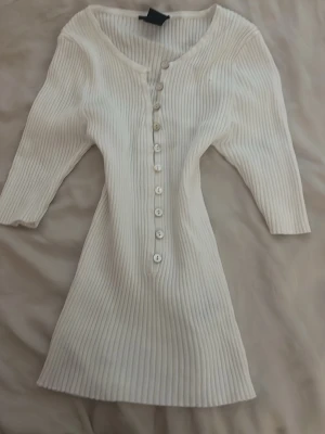 Vit ribbad lång topp med knappar - Snygg vit ribbad topp med knappar framtill. Toppen har en v-ringad halsringning och är tillverkad i ett stretchigt material som sitter bekvämt. Perfekt för dig som gillar enkel men vintage design.