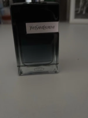 Yves Saint Laurent parfymflaska - Snygg parfymflaska från Yves Saint Laurent i glas med mörkblå till svart toning och silverfärgad metallplatta med logotyp på framsidan. Flaskan har en stilren, rektangulär form och svart kork. Perfekt för dig som gillar exklusiva dofter och elegant design. Använd några gånger men jag ska köpa en annan parfym så då säljer jag denna