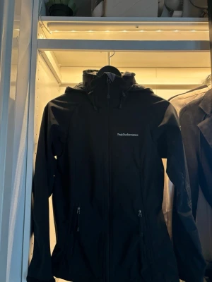 Svart vindjacka från Peak Performance - Snygg svart vindjacka från Peak Performance med huva och dragkedja framtill. Jackan har två fickor med dragkedja och diskret logga på bröstet. Perfekt för blåsiga dagar och enkel att matcha med olika outfits.
