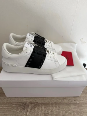 Valentino vita sneakers med nitar - Snygga vita sneakers från Valentino med svart detalj och svarta nitar på sidorna. Skorna är i läder och har klassisk låg modell med vita skosnören och diskret logga på plösen. Perfekta för dig som vill sticka ut med en edgy touch.