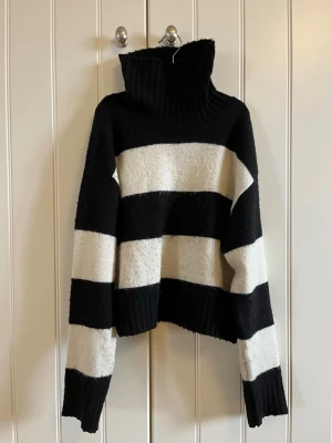 Svartvit randig polotröja Lindex - Stickad polotröja från Lindex med breda svarta och vita ränder. Tröjan har hög polokrage, långa ärmar och ribbstickade muddar. Perfekt för dig som gillar en avslappnad och trendig stil.