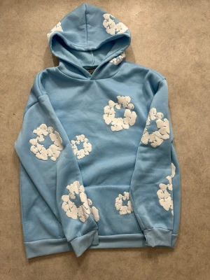 Blå hoodie från Denim Tears - Säljer en ljusblå hoodie från Denim Tears med vita blomliknande tryck över hela tröjan. Hoodien har en stor huva och klassisk känguruficka framtill. Tillverkad i mjukt och bekvämt material, perfekt för en chill och avslappnad stil.