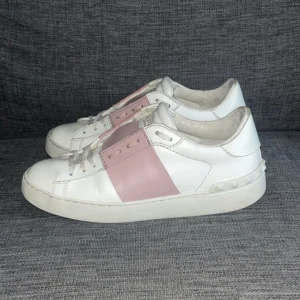 Valentino - Snygga Valentino Garavani sneakers i vitt skinn med bred rosa rem över vristen och ikoniska nitar på hälen. Klassisk rund tå, platt sula och snörning framtill. Perfekta för dig som vill ha en lyxig och trendig look.