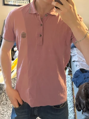 Rosa piké från DOLCE GABBANA - Säljer en rosa pikétröja från Dolce Gabbana, gråa detaljer på krage och ärmslut. Tröjan har korta ärmar, knappar framtill och en liten logga på bröstet. Perfekt för dig som vill ha en stilren och avslappnad look.