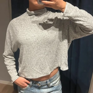 Grå croppad polotröja - Säljer en gråmelerad croppad polotröja med långa ärmar och lös passform. Tröjan har en hög krage och är tillverkad i ett mjukt, stretchigt material som känns skönt mot huden. Perfekt till jeans eller kjol för en avslappnad look.