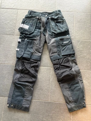 Mörkgrå arbetsbyxor Fristads - Säljer ett par mörkgrå arbetsbyxor från Fristads med flera praktiska fickor, förstärkta knän och detaljer i ljusgrått. Byxorna har hängfickor, verktygshållare och robusta sömmar, perfekta för dig som gillar funktionella plagg med streetkänsla. Använts 2 gånger.