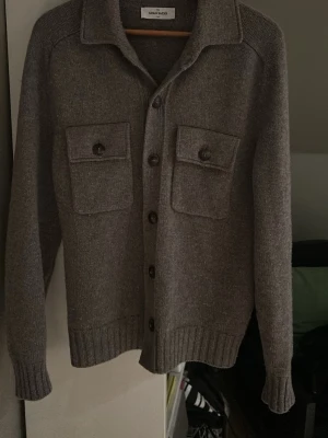 Grå kofta från Gran Sasso - Säljer en stilren grå kofta från Gran Sasso. Koftan har knäppning framtill, två stora bröstfickor med lock och knappar samt ribbade muddar vid ärmslut och nederkant. Perfekt för lager på lager och enkel att matcha med olika outfits.