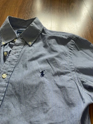 Randig skjorta från Ralph Lauren - Klassisk blåvit randig skjorta från Ralph Lauren med button-down krage och broderad logga på bröstet. Skjortan har långa ärmar, vita knappar och är tillverkad i mjuk bomull. Perfekt för en clean och stilren look. 43 B x74 L cm. 