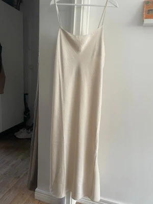 Champagnefärgad lång ”satin”klänning - Elegant långklänning i champagnefärgad satinliknande (polyester) material med tunna axelband och slits på ena sidan. Klänningen har en minimalistisk och stilren design som ger en lyxig känsla. Perfekt för dig som gillar en enkel men classy look. Ny med prislapp. Har lite ”rynkor” pga legat vikt men går nog att stryka bort. 