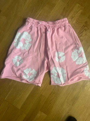 Rosa shorts från Denim Tears - Säljer ett par rosa shorts från Denim Tears med vita blommönster tryckta över hela plagget. Shortsen har resår i midjan och sidofickor. Tillverkade i mjukt bomullsmaterial, perfekta för en avslappnad och trendig stil.