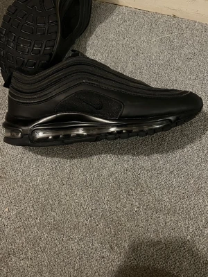 Nike Air Max 97 helsvart sneaker - Säljer ett par helsvarta Nike Air Max 97 sneakers med ikonisk vågig design och synlig Air-sula runt hela skon. Ovandelen är i en mix av mesh och syntetmaterial för en clean look. Perfekta för dig som gillar streetwear och vill ha något stilrent.